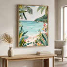 A framed tropical beach art print hangs on a light beige wall above a wooden console table. The artwork features lush green palm leaves, a sandy beach, turquoise ocean water, and colorful tropical flowers.