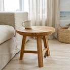 A round wooden side table with three legs and a small attached storage loop, styled in a bright living room next to a beige sofa with a white mug on top.