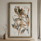 A framed botanical art print featuring abstract watercolor leaves and berries in earthy brown, beige, and gray tones, displayed on a light wood console table next to a small vase with dried branches.