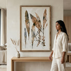 A woman in a white linen outfit stands beside a framed botanical wall art print in a warm, modern living room. The print features watercolor-style leaves and stems in earthy brown, beige, and muted blue-gray tones on a light background, enclosed in a natural wood frame.