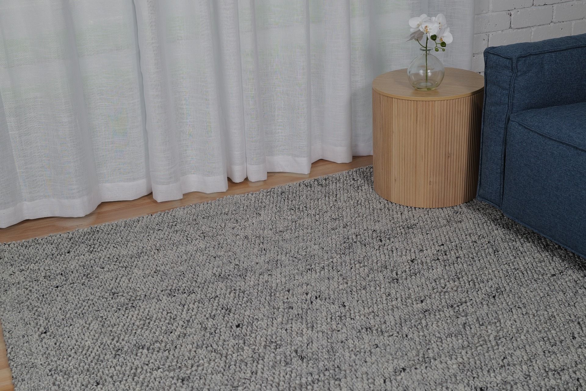 Living room with a gray textured rug, white curtains, a wooden side table, and a blue sofa.