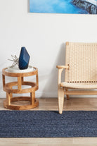 Wooden side table with a blue vase and plant, next to a woven chair on a blue rug.
