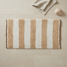 A rectangular bath mat with vertical tan and off-white stripes, laid flat on beige tiled flooring with a folded beige towel and a small seashell nearby.