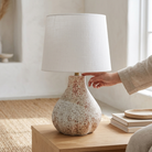 A table lamp with a textured, speckled ceramic base and a white linen drum shade, shown on a wooden table in a bright room with a person's hand reaching toward the lamp.