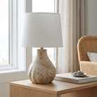 A table lamp with a textured, speckled beige ceramic base and a white fabric drum shade, styled on a light wood side table in a bright room next to a window.