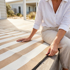 A person sitting on a wooden deck outdoors, wearing a white button-up shirt and light beige pants, with a striped woven rug in beige and off-white extending across the deck.