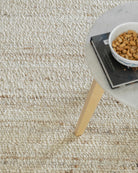 Close-up of a textured beige carpet with a small table and bowl of cat food in the corner.