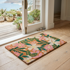 A rectangular coir doormat with a colorful floral pattern in pink, green, cream, and orange, placed on a light wood floor in front of an open white door leading to a deck and ocean view.