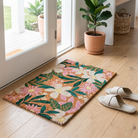 A rectangular coir doormat with a colorful tropical floral pattern lies on a light wood floor in a bright entryway. A potted plant, woven basket, and a pair of off-white slide sandals are nearby.