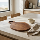 A round wooden serving board with a loop handle sits on a light stone kitchen island, alongside a speckled bowl and a beige linen napkin. The scene is set in a bright, modern kitchen with natural light coming from a window.