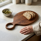 A dark brown wooden serving board with a loop handle sits on a light stone countertop. Slices of whole wheat bread rest on the board, and a hand in a beige linen sleeve rests beside it.