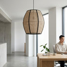 A large woven rattan pendant light with black metal framing hangs in a modern, bright office space. In the background, a man in a light beige shirt sits at a wooden desk next to a small potted plant.