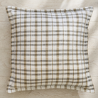 A square throw pillow with a brown and off-white plaid pattern, resting on a light wood floor.