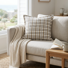 A beige and brown plaid throw pillow displayed on a light gray sofa in a bright, modern living room setting with a cream knit blanket, a wooden side table, and an open book.