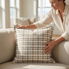A woman in a cream linen shirt sits on a beige sofa, holding a square throw pillow with a brown and beige plaid pattern in a brightly lit living room.