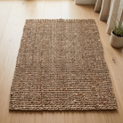 A rectangular natural jute rug with a textured, ribbed woven pattern, shown on a light-colored wooden floor next to a curtain and a small potted plant.