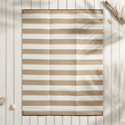A rectangular beach towel with horizontal tan and white stripes, laid flat on a white wooden plank surface with decorative seashells and driftwood.