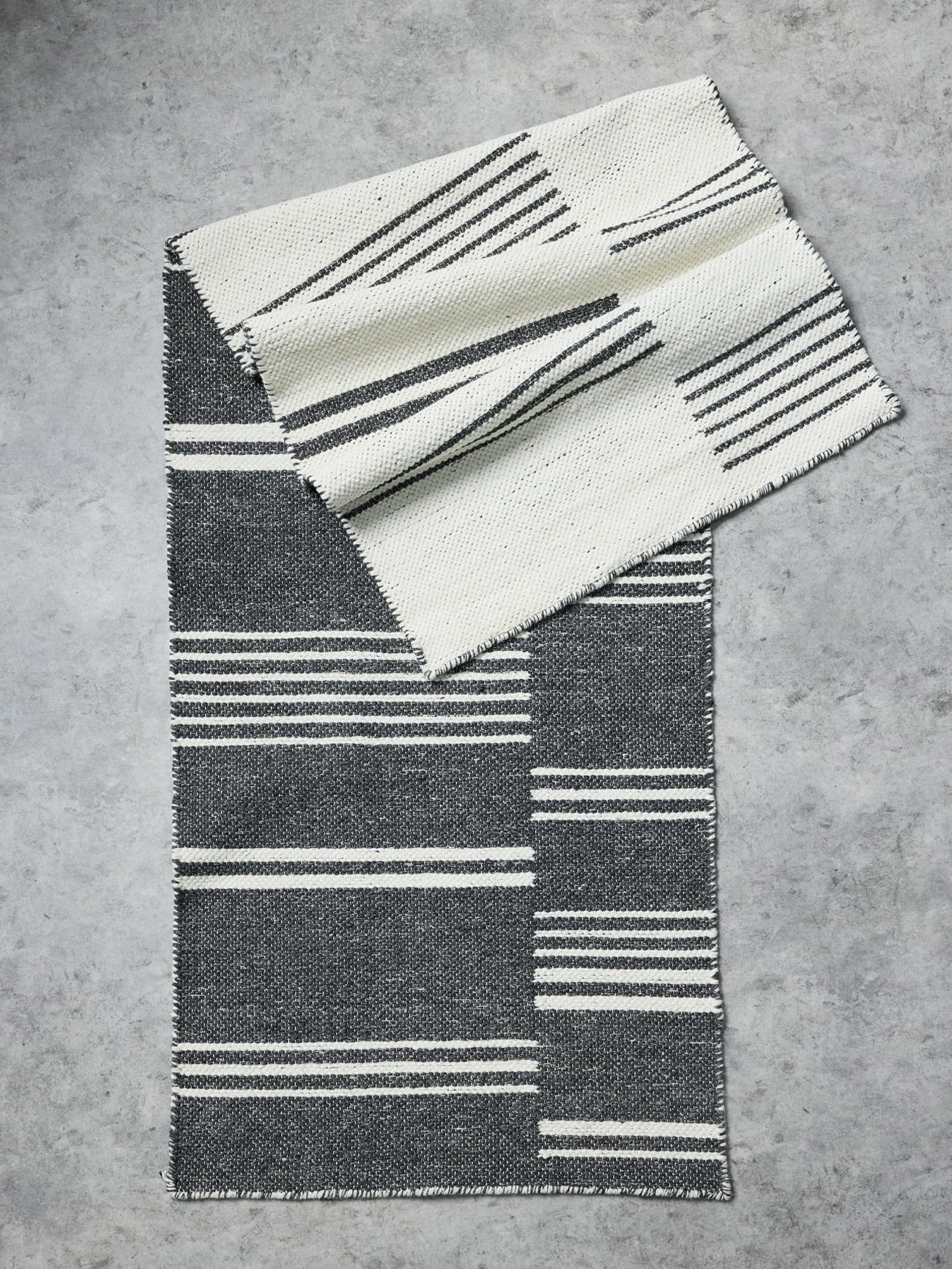 A folded woven textile with a black and off-white striped geometric pattern, displayed on a textured gray concrete surface.