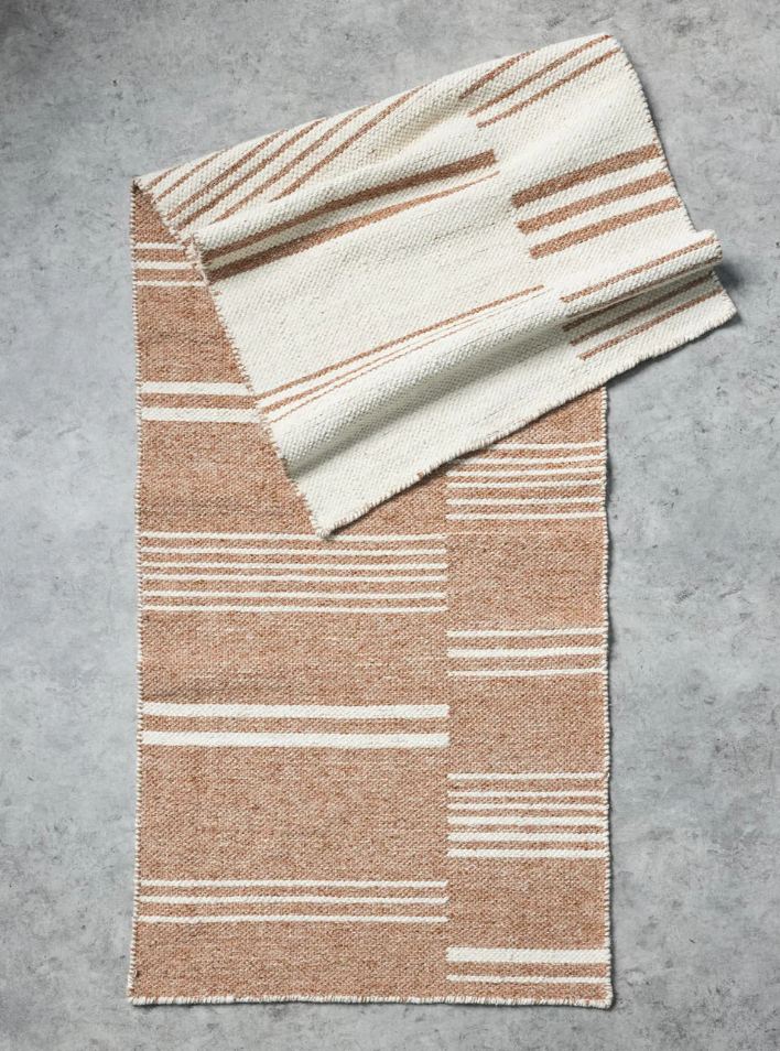 A woven rectangular throw blanket with a striped pattern in warm tan and cream, folded diagonally on a textured gray concrete surface.