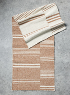 A woven rectangular throw blanket with a striped pattern in warm tan and cream, folded diagonally on a textured gray concrete surface.