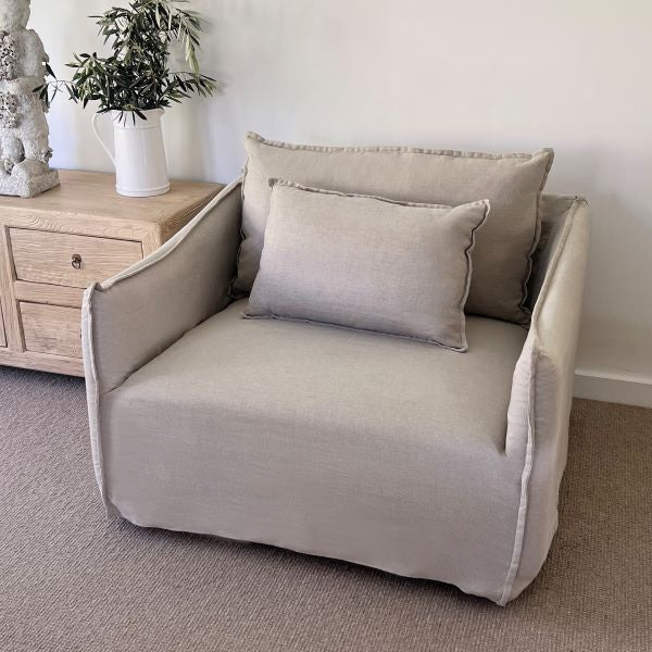 Beige armchair with cushions in a room setting with a wooden side table and plant.