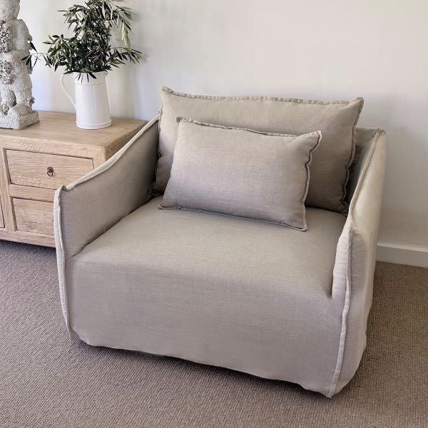 Beige armchair with cushions in a room setting with a wooden side table and plant.