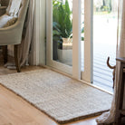 Beige woven rug on a wooden floor in a room with a sliding glass door and plant.