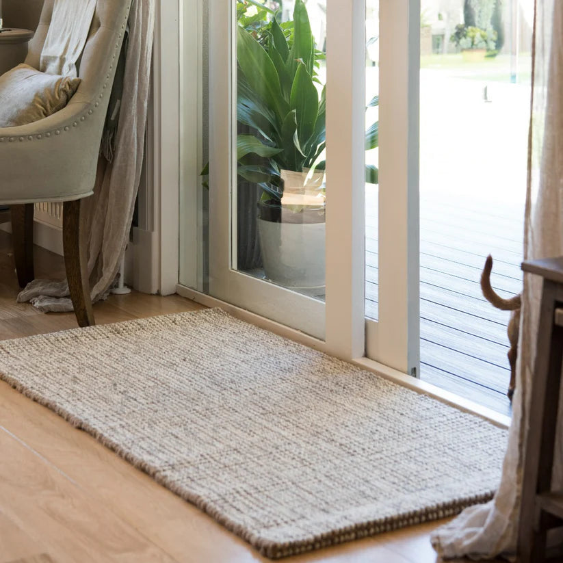 Beige woven rug on a wooden floor in a room with a sliding glass door and plant.