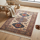 A rectangular area rug with a traditional geometric pattern in beige, blue, red, and brown, styled on a light wood floor in a bright room next to a wooden armchair and sandals.