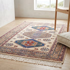A rectangular area rug with a traditional geometric pattern in cream, blue, red, and brown tones, styled on a light wood floor in a bright room next to a wooden chair and a cushion.