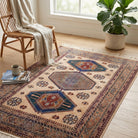 A rectangular area rug with a traditional geometric pattern in beige, blue, red, and brown tones, styled on a light wood floor in a bright room next to a wicker chair with a cream throw blanket and a potted fiddle-leaf fig plant.