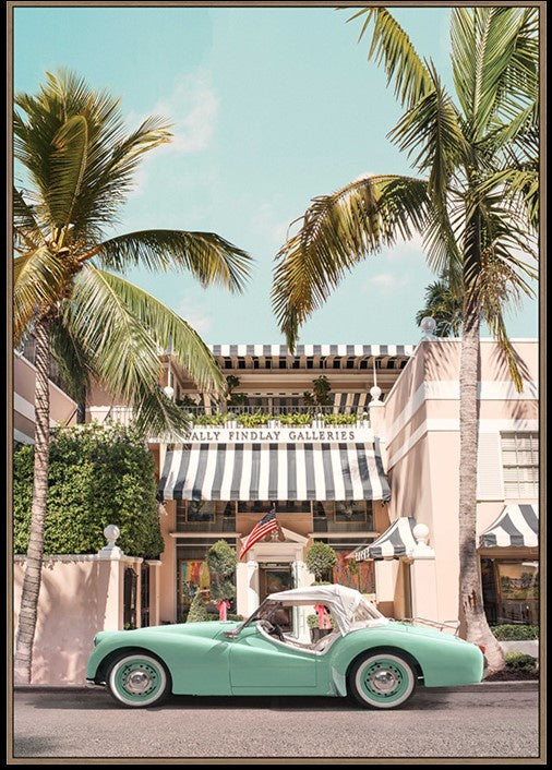 A framed art print depicting a retro mint green convertible car parked on a street in front of a building with a black and white striped awning, framed by tall palm trees under a light blue sky.