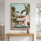 A framed vertical art print depicting a retro turquoise convertible car parked in front of a pink beachside building with palm trees, displayed on a white wall above a light wood console table in a modern living room setting.