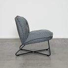 A modern gray upholstered accent chair with a black metal X-frame base, shown in profile against a white wall and concrete floor.