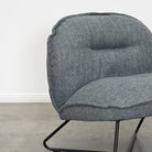 A close-up of a modern upholstered accent chair with a textured gray fabric and a black metal wire frame, set against a plain white wall and concrete floor.