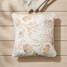A square decorative throw pillow with a beige and off-white base, featuring a soft botanical and seashell pattern in muted peach, tan, and pale blue, styled on a light wood plank surface.