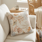 A square decorative throw pillow with a nautical shell and coral pattern in muted beige, tan, and soft blue-gray tones, styled on a cream-colored sofa in a bright living room setting.
