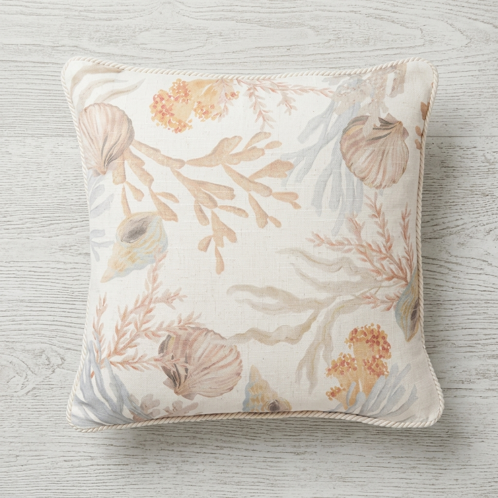 A square decorative throw pillow with a nautical design featuring seashells, coral, and seaweed in muted beige, peach, and light blue-gray tones on an off-white background, resting on a light gray wood plank surface.