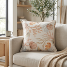 A square decorative throw pillow with a beige and cream background and an ocean-themed print of seashells, coral, and jellyfish, styled on a light beige armchair in a cozy living room setting.