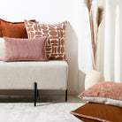 A styled interior scene with a light upholstered bench or sofa holding several decorative throw pillows in rust, dusty pink, and cream-and-brown patterned fabric. A tall white vase with dried grass stands against a white wall, and part of another cushion is visible in the foreground on a light rug.