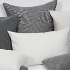 A styled arrangement of multiple decorative throw pillows in neutral tones, including a prominent white ribbed pillow in the foreground, a black-and-white geometric patterned pillow behind it, and several gray and off-white pillows.