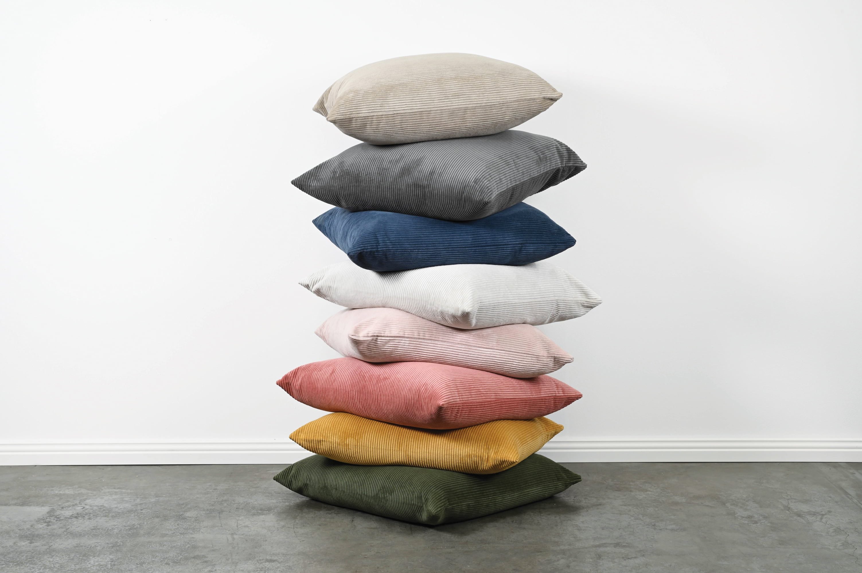 A stack of nine throw pillows in various colors, including beige, gray, navy, white, light pink, dusty pink, mustard yellow, and olive green, arranged on a gray floor against a white wall.