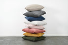 A stack of nine throw pillows in various colors, including beige, gray, navy, white, light pink, dusty pink, mustard yellow, and olive green, arranged on a gray floor against a white wall.