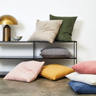A collection of textured throw pillows in various colors including beige, olive green, gray, pink, yellow, white, and blue, styled on a black metal shelf and the floor against a white wall.