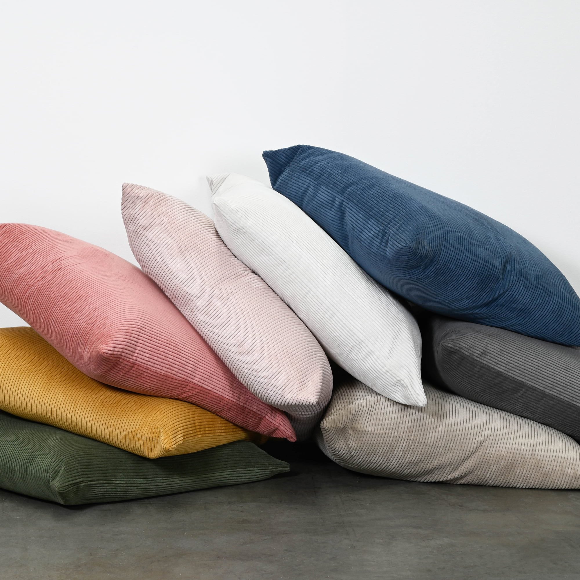 A stack of six ribbed corduroy throw pillows in various colors including navy blue, dusty pink, light pink, white, olive green, mustard yellow, and gray, arranged on a dark gray floor against a white wall.