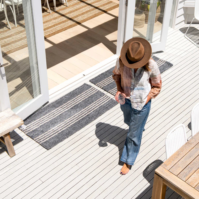 Person walking on a wooden deck with a rug, wearing a hat and plaid shirt.