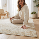 A woman in a cream knit sweater and matching pants sits cross-legged on a large beige textured area rug in a bright, modern living room with light wood floors and plants.