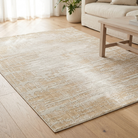 A rectangular area rug with a distressed abstract pattern in beige and cream tones, shown on a light wood floor in a bright living room setting.