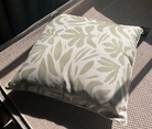 A square throw pillow with a sage green tropical leaf pattern on a cream background, resting on a woven outdoor patio chair in direct sunlight.