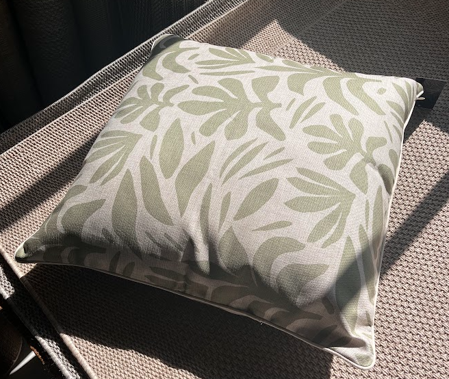 A square throw pillow with a sage green tropical leaf pattern on a cream background, resting on a woven outdoor patio chair in direct sunlight.